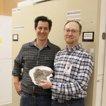 Two men stand amongst museum collections compactors and hold a small fossil