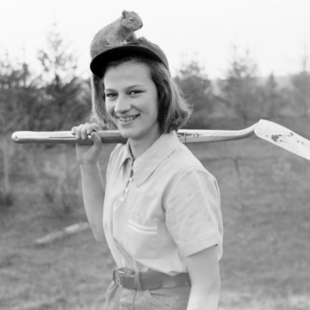 Woman in a black and white photo with a shovel over her shoulder smiles at the camera. A squirrel sits on top of her head.