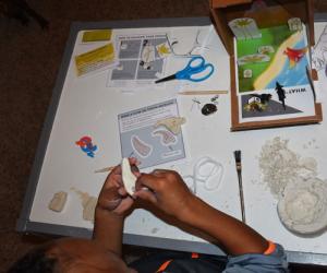 a child makes a clay tooth necklace in the fossil finder fun pack box from the Burke Museum